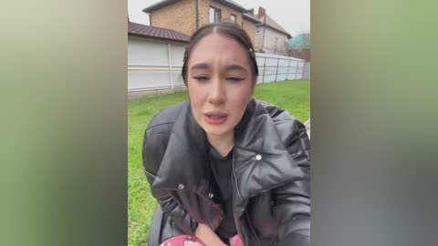 A video shows a young Asian woman with long dark hair in a black leather jacket, sitting on a grassy area, looking distressed. Houses and a white fence are in the background.