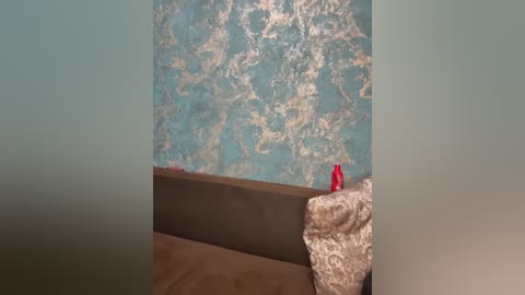 Video of a bedroom corner with textured turquoise and gold wallpaper, a brown wooden bed, beige and gold lace bedspread, and a red toy on the pillow.