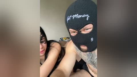 Video: A tattooed man in a black balaclava with \"$Money\" written on it performs oral sex on a woman wearing a black lace mask and a black bra, in a dimly lit room with beige walls.