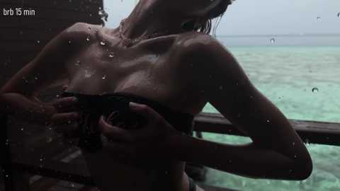 Video of a topless woman with wet hair, small breasts, and a slim physique, standing on a wooden deck overlooking a turquoise ocean. She holds a black object close to her chest.