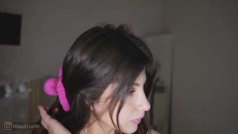 Video of a young woman with long dark hair, wearing a bright pink hair tie, focused on combing her hair in a dimly lit room with a white wall and a partially visible window.