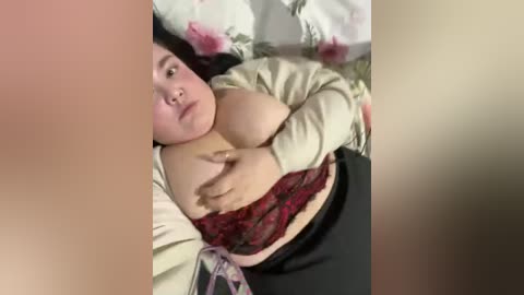 Video of an overweight Asian woman with large breasts, partially covered by a red lace bra and beige cardigan, lying on a bed with floral-patterned sheets.