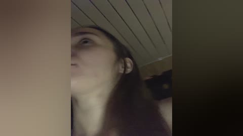 Video of a young woman with fair skin and long brown hair, wearing a black top, standing in a dimly lit room with a white ceiling.