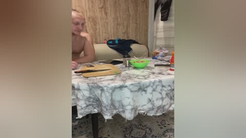 A blurry video of a nude blonde woman, seated at a cluttered table, in a dimly lit, rustic room with wooden walls and a beige couch.