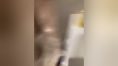 Blurry video of a person in a beige shirt and light-colored pants, standing in front of a white fridge with a yellow box on top, partially obscured by a white pillar.