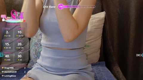 A video captures a light-skinned woman in a light blue tank top and skirt, sitting on a bed with a patterned pillow and dark curtains. The image includes a virtual reality overlay with stats and a pink circle on the top right.