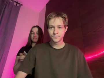 Video of a young man in a dark room with pink lighting, wearing a black shirt, and a woman with long hair and a black shirt, holding a camera.