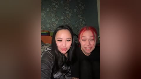 Video of two Asian women with long black hair and vibrant red hair, smiling playfully, in a dimly lit room with floral wallpaper and colorful storage boxes.