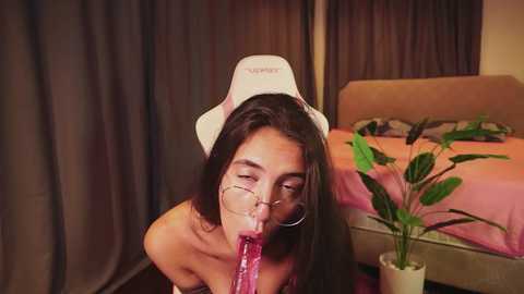 A video shows a naked Asian woman with long black hair, wearing a white nurse cap, sucking on a large, pink dildo. She's in a dimly lit bedroom with a bed, curtains, and a potted plant.