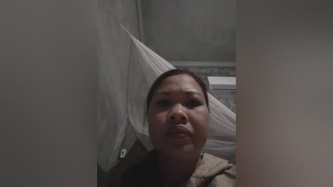 Video of a distraught, middle-aged Asian woman with a bandage around her head, lying on a bed in a dimly lit, gray-walled room with a white door and a white cabinet.