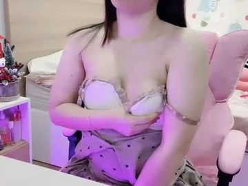 Video of a light-skinned woman with long black hair, wearing a strapless white bra, sitting in a pink gaming chair. Background includes a bed and desk with scattered objects.