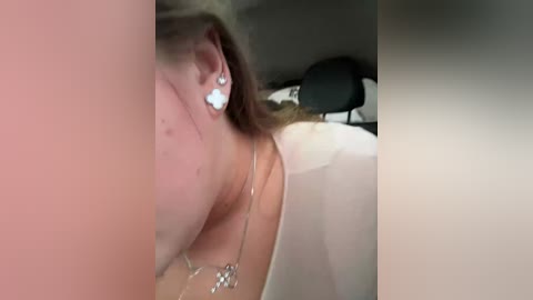 Video of a woman with light skin, brown hair, wearing a white shirt, silver necklace, and stud earrings, lying on a bed with a black headrest, blurry background.