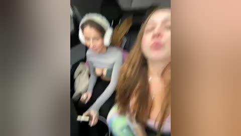 A blurry video captures two young women in a car, one with headphones, the other kissing, wearing casual outfits.
