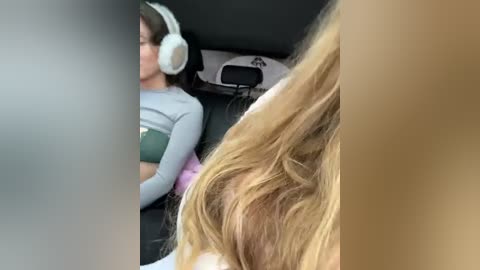 A video of a woman with long, blonde hair wearing headphones, sitting in a car with a gray shirt.