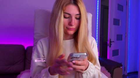 Video of a young woman with long blonde hair, wearing a white sweatshirt, using a silver smartphone, seated on a gray couch in a dimly lit room with purple and blue LED lighting.