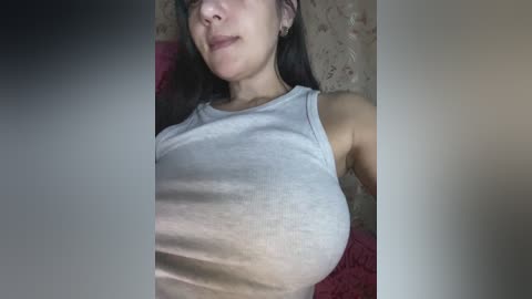 Video of a young woman with medium skin tone and long, straight dark hair, wearing a tight, light gray tank top that accentuates her medium-sized breasts, against a textured, beige wall background.