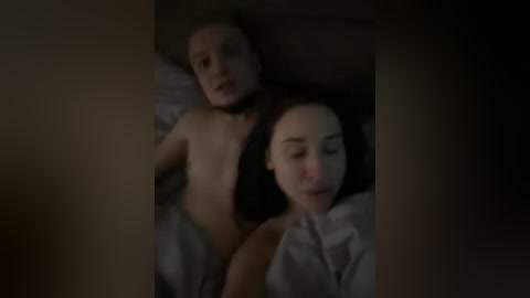 A dimly-lit video of two young women with fair skin and dark hair lying side by side on a bed, partially covered by white sheets, both topless and looking relaxed.