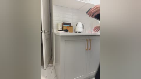 Video of a modern kitchen sink area with white cabinets, a white kettle, and a person's hand washing dishes, partially visible. The background features white subway tiles and a door on the left.