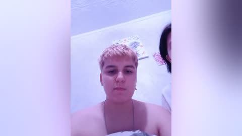 Video of a young woman with short, dyed pink hair, wearing a white top, standing in a small, white bathroom with a circular mirror and colorful towels on the counter.