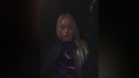 Video of a solemn blonde woman in a dark, shadowy setting, wearing a purple dress, her face partially illuminated, with a somber expression.