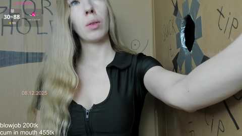 A video of a blonde woman with fair skin wearing a black zipped shirt, captured in a room with a cardboard cutout of a man and a \"FOR SALE\" sign.