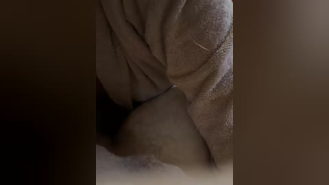 A dimly-lit video captures the close-up of a person's lower abdomen and upper thigh. The skin is light-toned, with visible folds and creases. The individual is wearing a beige sweater with a textured, ribbed pattern. The background is blurred, emphasizing the subject's body.