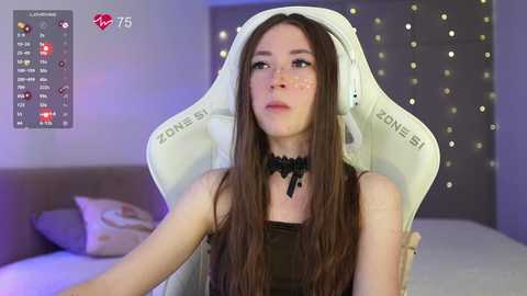 Video of a young woman with long brown hair, fair skin, and a black choker, sitting in a white gaming chair, in a dimly lit bedroom with a bed and string lights.
