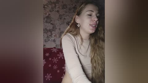 Video of a young, light-skinned woman with long, wavy blonde hair, wearing a beige long-sleeve top, sitting on a red cushion with white snowflake pattern. She appears joyful, eyes closed, lips parted. Background features floral wallpaper.