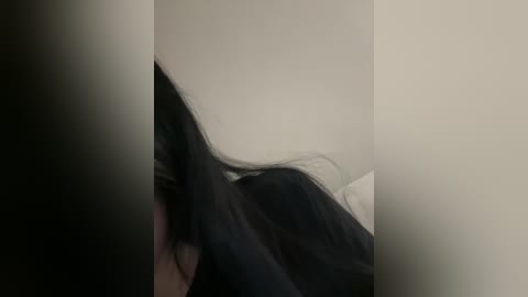 A video shows a person with long, straight black hair partially obscuring their face, lying on a bed with white sheets. The background is a plain, light beige wall, creating a simple, minimalist setting.