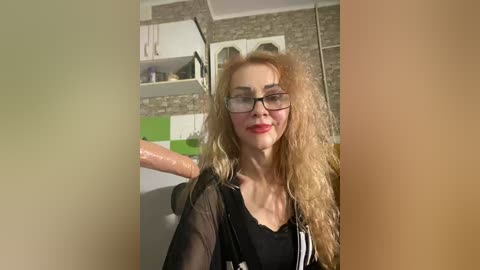 A video of a blonde woman with curly hair, wearing glasses and a black top, in a modern kitchen with green and white tiles.