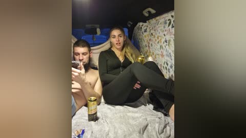 Video of a shirtless man and a woman in black clothing, sitting on a bed with a beer can and a glass in hand, in a dimly lit room with colorful wall art.