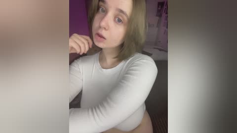 A young woman with fair skin, blue eyes, and shoulder-length blonde hair, wearing a white long-sleeve shirt, poses indoors with a neutral expression.