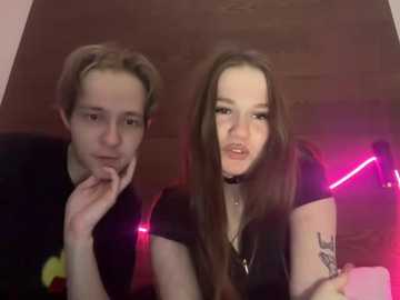 Video of a young couple, a boy with short hair and a girl with long hair, both with pale skin, indoors with pink neon lights, holding a phone and smiling.