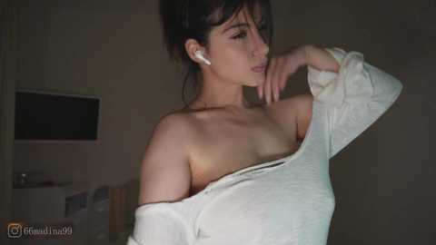 Video of a young woman with light skin, dark hair, wearing a white off-shoulder sweater, standing in a dimly lit room, looking contemplative.