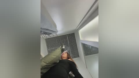 Video of a woman with long hair, wearing a black sweater, lying on her back in a modern, minimalist room with white walls, black-tiled floor, and a grey couch.