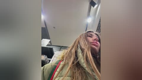 Video of a young woman with long, wet, blonde hair, wearing a green jacket, sitting in a car with a blurry, bright light in the background.