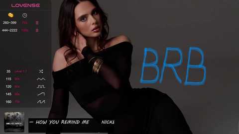 Video of a woman with long, dark hair, wearing a sheer black off-shoulder top and black dress, leaning against a gray background. \"BBB\" is prominently displayed.