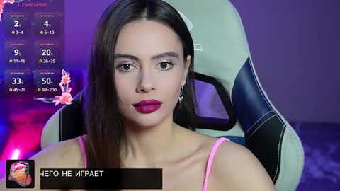 Video of a woman with long dark hair, wearing pink spaghetti straps, sitting in a gaming chair, surrounded by a digital screen displaying a calendar and statistics.