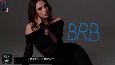 Video of a woman with long dark hair, wearing a sheer black off-shoulder dress, sitting against a dark gray backdrop with \"BBB\" text.