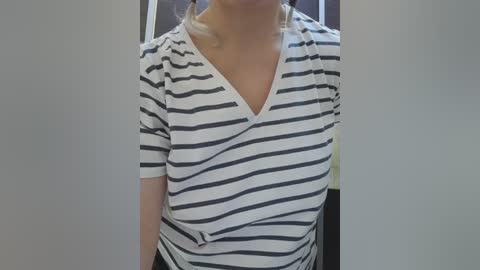 A video of a woman wearing a white and black striped V-neck top, with a slight twist to the waistband, partially visible blonde hair, and a blurred background.