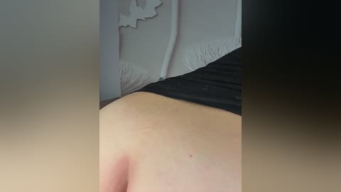 A close-up video of a person's upper back and lower neck, showing light skin, a black shirt, and a white decorative wall with a floral pattern.