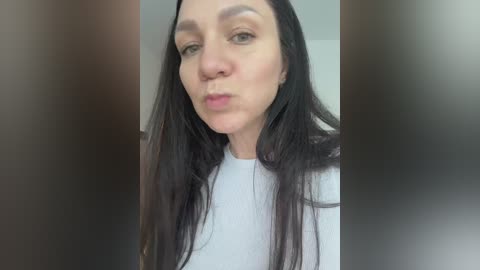 A video of a woman with long, straight black hair, fair skin, and light blue eyes, wearing a white shirt. She has a neutral expression. The image is slightly blurry with a dark border.