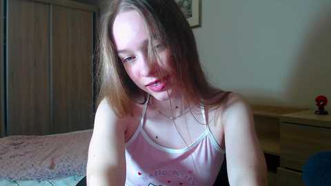 A video of a young, fair-skinned, blonde girl in a white tank top, smiling slightly, in a bedroom with a wooden wardrobe and a bed with pink bedding.