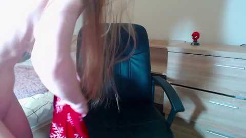 Video of a woman with long brown hair, seated in a black office chair, wearing a red floral dress, in a modern bedroom with light wooden furniture and a red lamp.