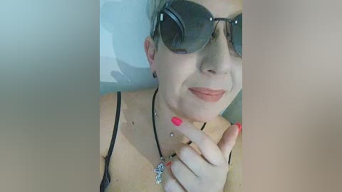 Video of a middle-aged woman with short blonde hair, wearing dark sunglasses, a black spaghetti strap top, and red nail polish, with a necklace, and resting on a white pillow.