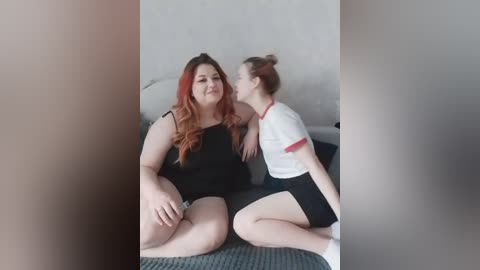 Video of two plus-sized women with fair skin, one with long red hair in a black dress, the other with brown hair in a white and red sports top, kissing on a couch in a plain, grey-walled room.