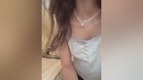 A blurred video of a woman with long, dark hair, wearing a white blouse and silver necklace, standing near a beige countertop with a metal bowl containing colorful objects.