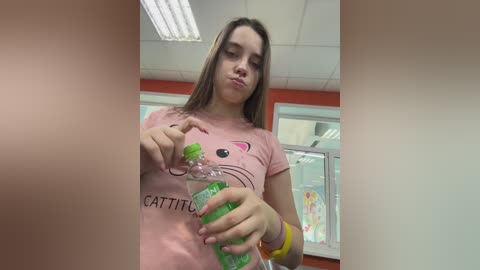 Video of a young Caucasian woman with light skin, straight brown hair, and a pouty face, wearing a pink t-shirt with cat ears and a yellow bracelet, pouring green drink into a glass in a brightly lit room.