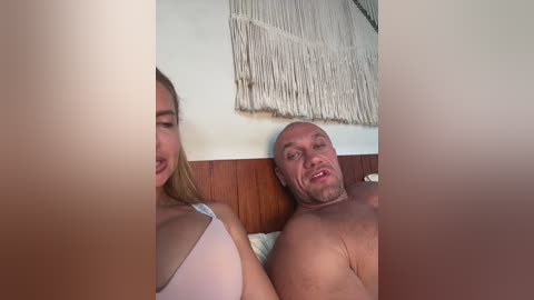 Video of a blonde woman and bald, muscular man lying shirtless on a bed, background showing a macrame wall hanging.