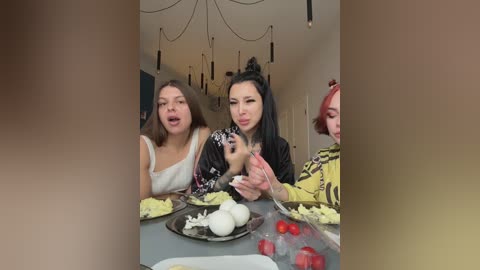Video of three women, one Asian with long black hair, one with brown hair, and one with red hair, seated at a table with food, including eggs and tomatoes, in a modern, dimly lit room.